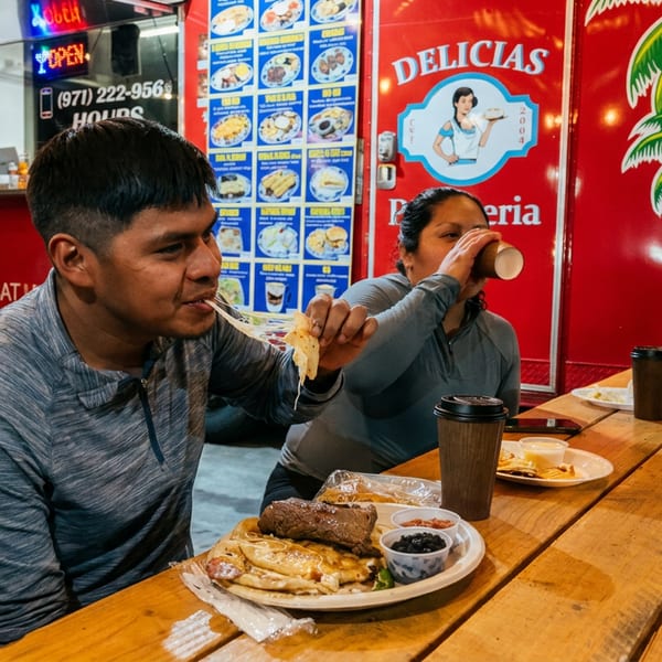 Customers enjoy cheesy food and drinks at a wooden picnic table outside a red "DELICIAS" food truck.