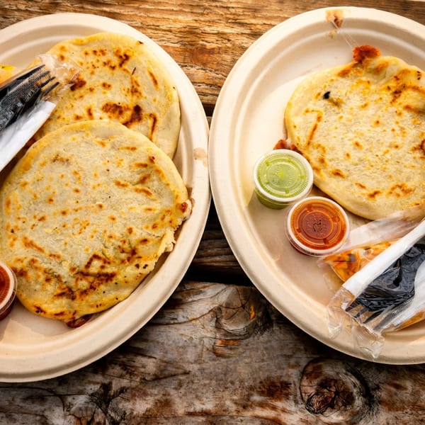 Two plates of pupusas with sauces and plastic cutlery on a rustic wooden table.