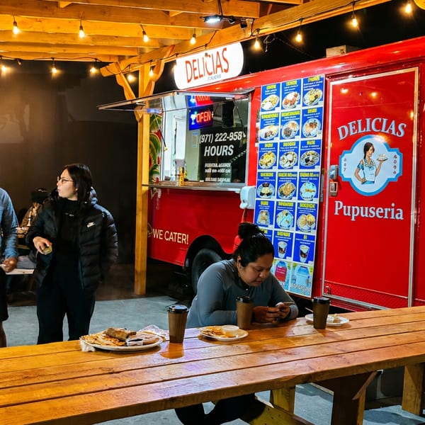 Customers enjoy food from the red Delicias Pupuseria food truck at illuminated outdoor picnic tables at night.